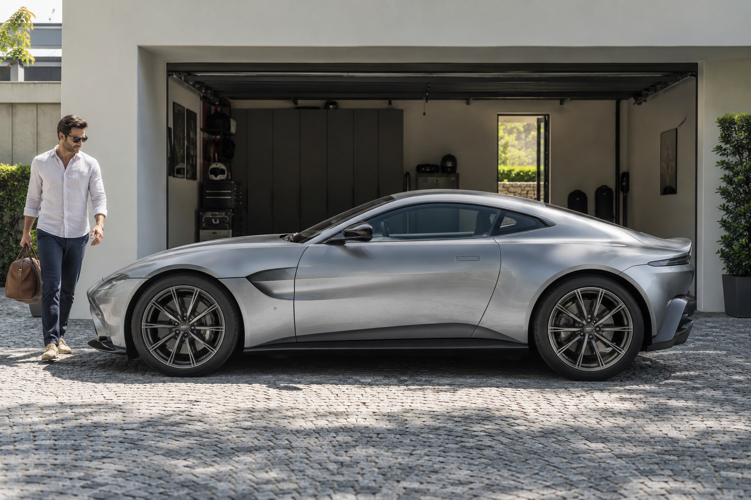 Aston Martin Kfz-Versicherung: Individualausstattung und Nutzung eines Vantage, DB12 oder DBX nachvollziehbar absichern. Silbernes Sportcoupé mit Mann vor einer Garage: Aston Martin Autoversicherung