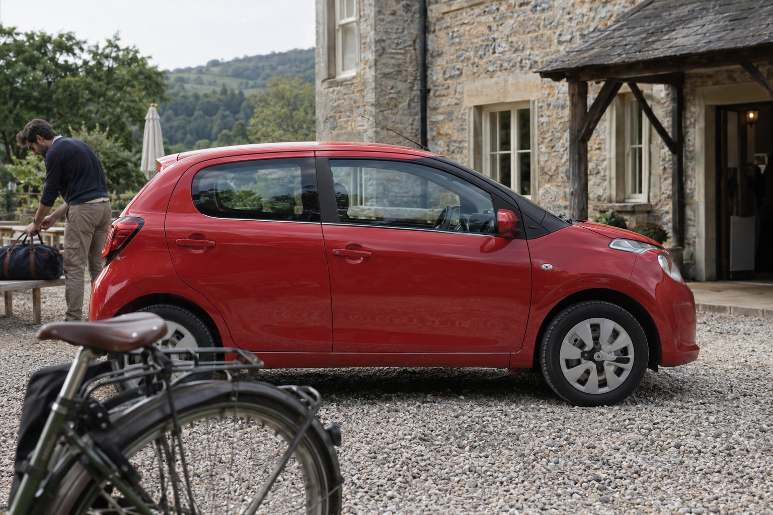 Citroën C1: Beim C1 Airscape spielt das textile Dach eine Rolle, wenn Kaskoleistungen und Selbstbeteiligung verglichen werden. Roter Citroën C1 auf einem Kieshof mit Reisetasche und Holzbank