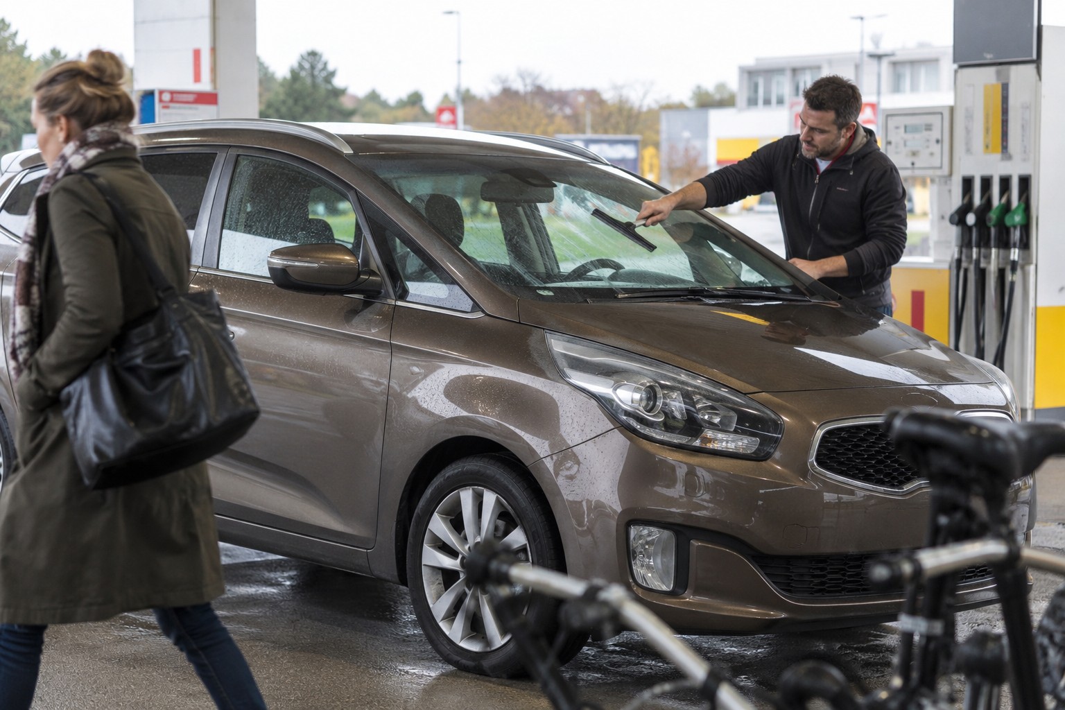 Kia Carens: Carens-Generationen und Sitzkonfigurationen gehören zur Fahrzeugprüfung, bevor der Familien-Van versichert wird. Bronzefarbener Kia Carens unter einem Tankstellendach beim Reinigen der Windschutzscheibe