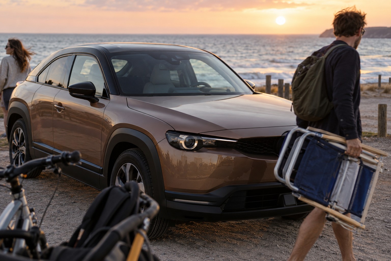 Mazda MX-30: Beim MX-30 R-EV arbeitet der Wankelmotor als Stromerzeuger; die reine Elektroausführung besitzt diese Technik nicht. Bronzefarbener Mazda MX-30 auf einem Parkplatz an der Küste mit Strandstühlen