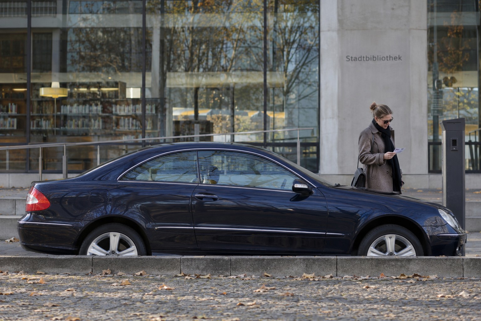 Mercedes-Benz CLK: Beim CLK Cabriolet unterscheiden sich Verdeckschäden von normalem Verschleiß des Dachsystems. Dunkelblauer Mercedes-Benz CLK vor einer Bibliothek neben einer Person mit Parkschein