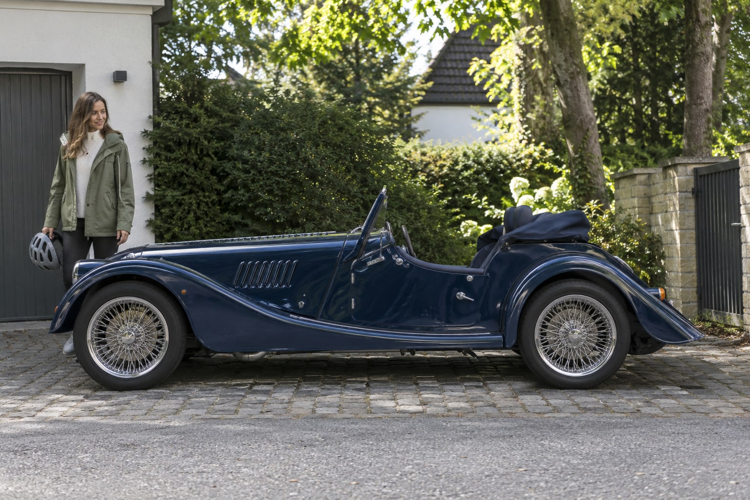 Morgan Kfz-Versicherung: moderne Roadster und Super-3-Dreirad mit eigenen Fahrzeugdaten unterscheiden. Blauer klassisch gestalteter offener Roadster mit Frau vor einem Haus: Morgan Autoversicherung
