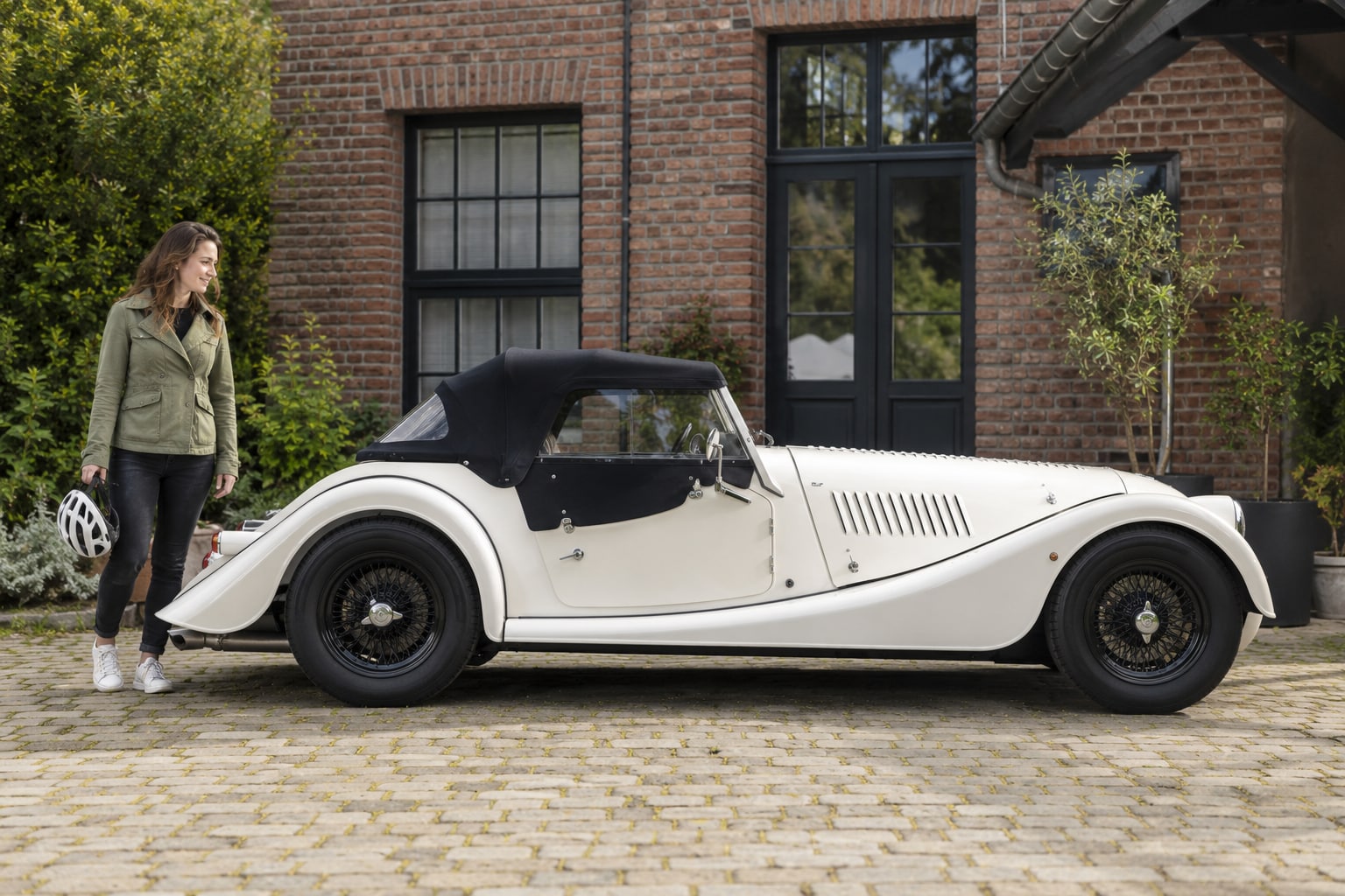 Morgan Plus Four Kfz-Versicherung: moderne Roadstergeneration und individuelle Ausführung passend zuordnen. Weißer offener Roadster mit Frau vor einem Backsteingebäude: Morgan Plus Four Kfz-Versicherung