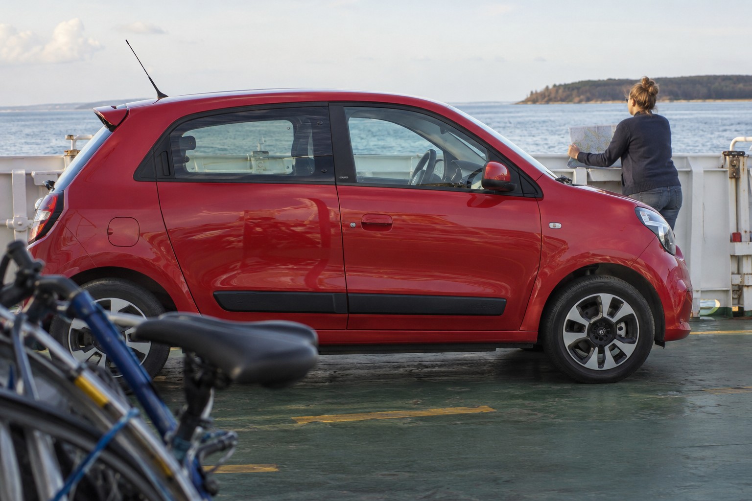 Renault Twingo: Twingo III mit Heckmotor und ältere Frontmotor-Baureihen sind unterschiedliche Versicherungsvarianten. Roter Renault Twingo auf einem Fährdeck neben einer Person mit Straßenkarte