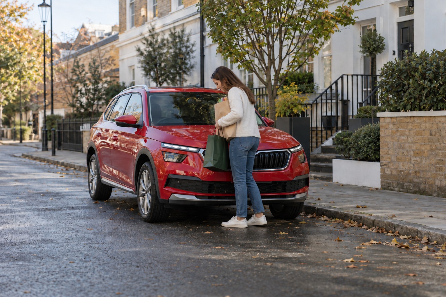 Škoda Kamiq Autoversicherung und Fahrerprofil Roter Škoda Kamiq in einer Wohnstraße; eine Frau hebt Einkaufstaschen