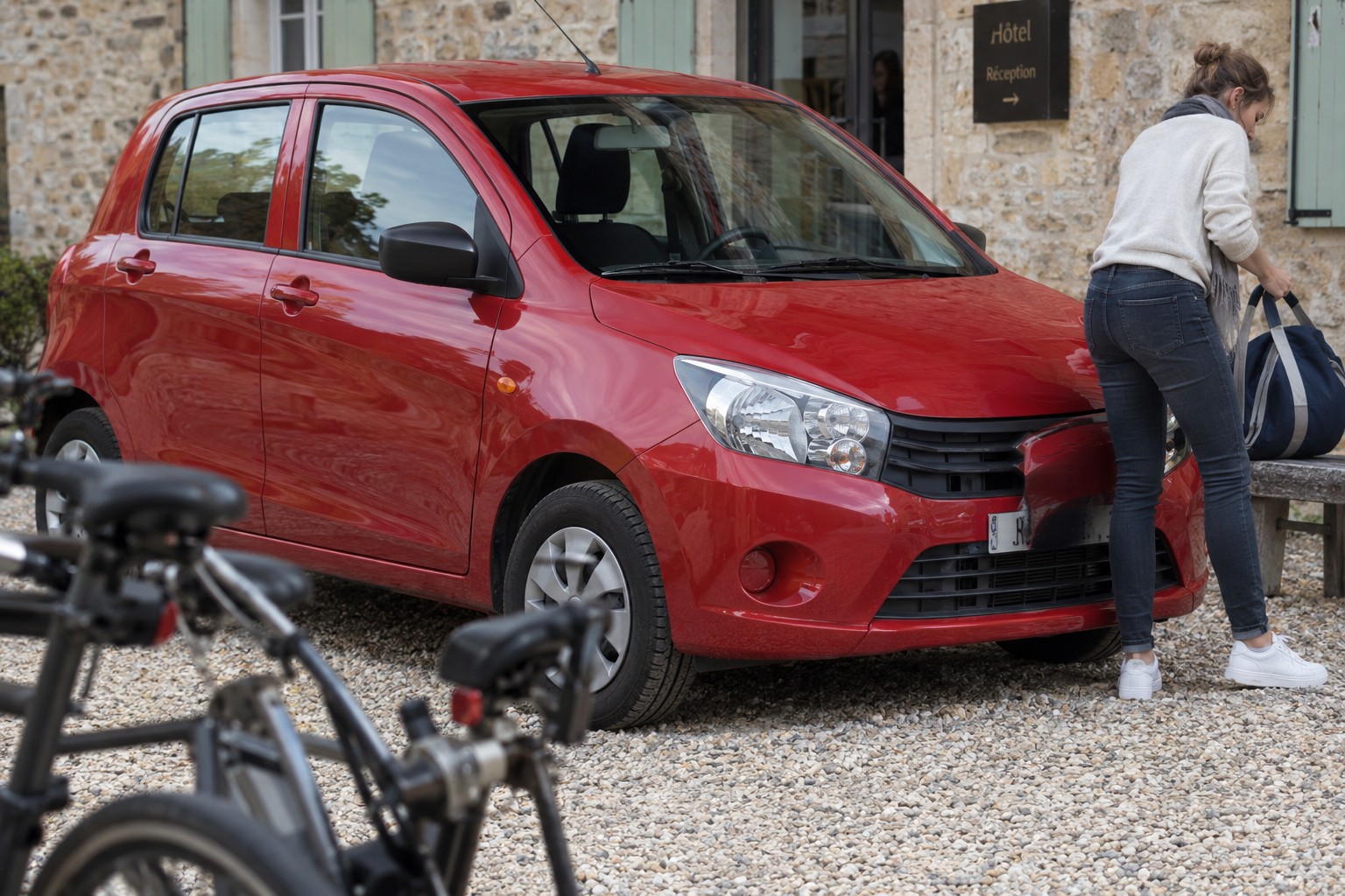 Suzuki Celerio: Für den Celerio ist die zugelassene Marktversion wichtiger als die Bezeichnung eines späteren ausländischen Modells. Roter Suzuki Celerio auf einem Kieshof mit Reisetasche und Holzbank
