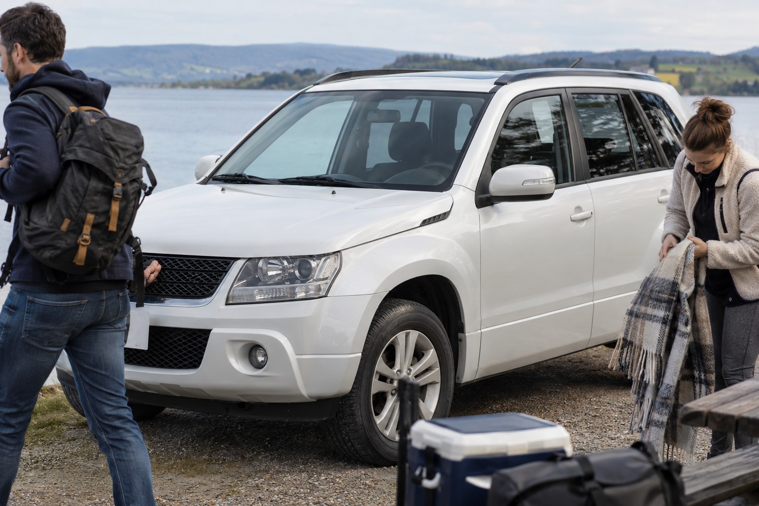 Weißer Suzuki Grand Vitara auf einem Seeparkplatz neben einer Person mit Picknickdecke