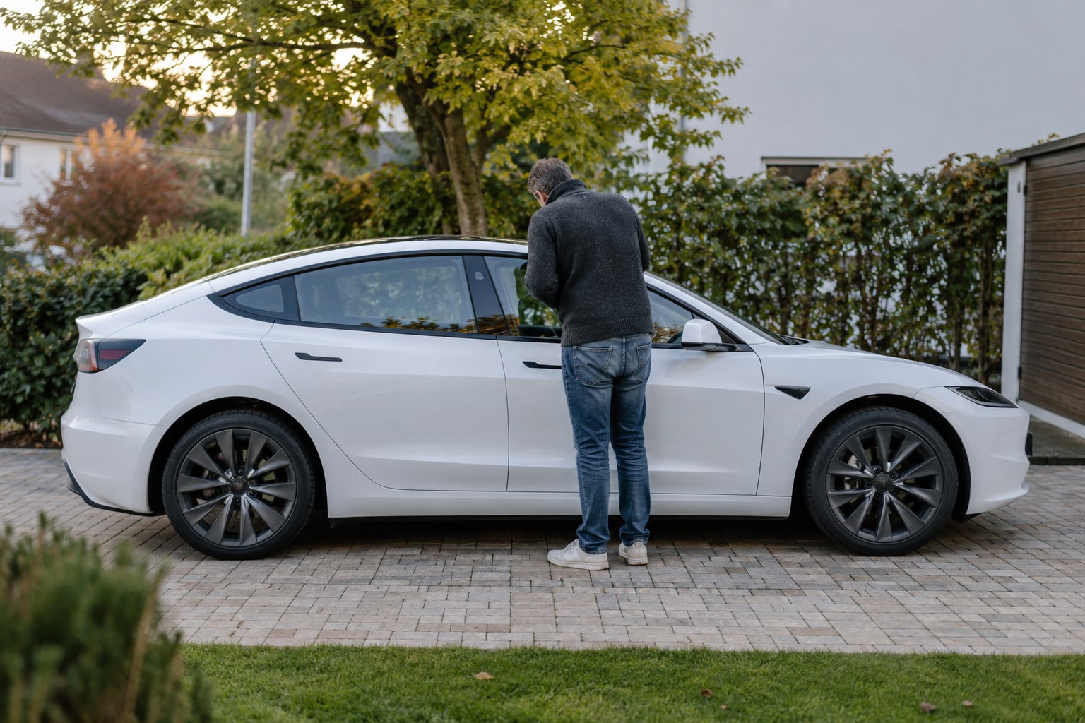 Tesla Autoversicherung: Elektroversion mit Akku-, Glas- und Werkstattleistungen vergleichen. Weiße Elektrolimousine mit Mann vor einem Garten: Tesla Autoversicherung