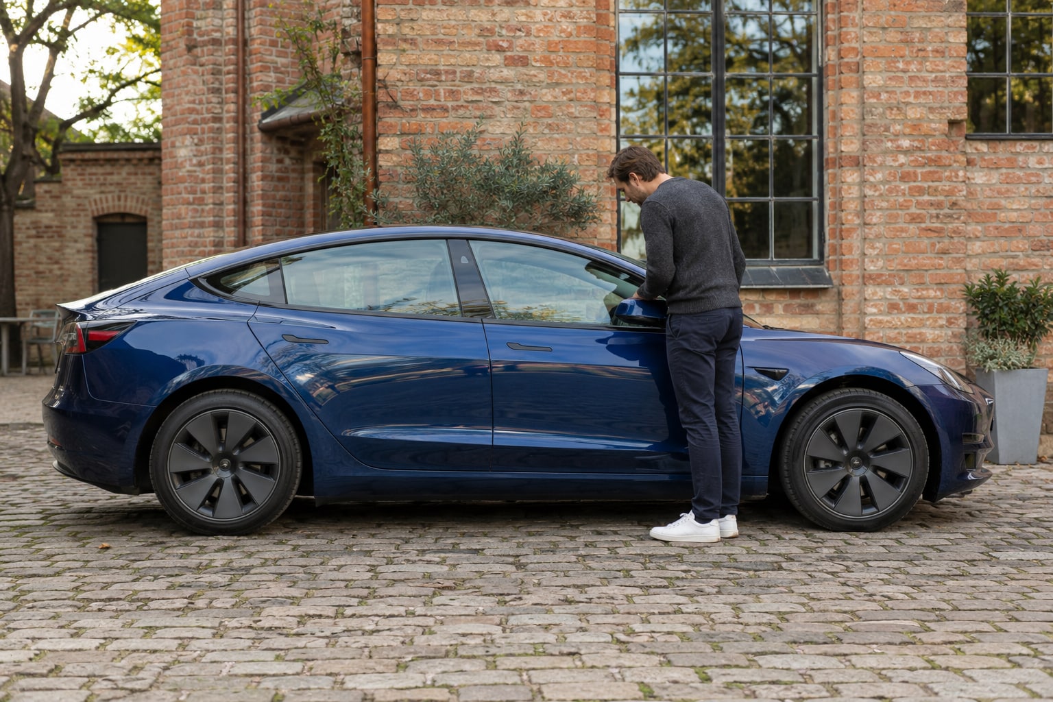 Tesla Model 3 Kfz-Versicherung: Limousinenversion mit Glas-, Kamera- und Akku-Schutz vergleichen. Blaue Elektrolimousine mit Mann vor einem Backsteingebäude: Tesla Model 3 Kfz-Versicherung