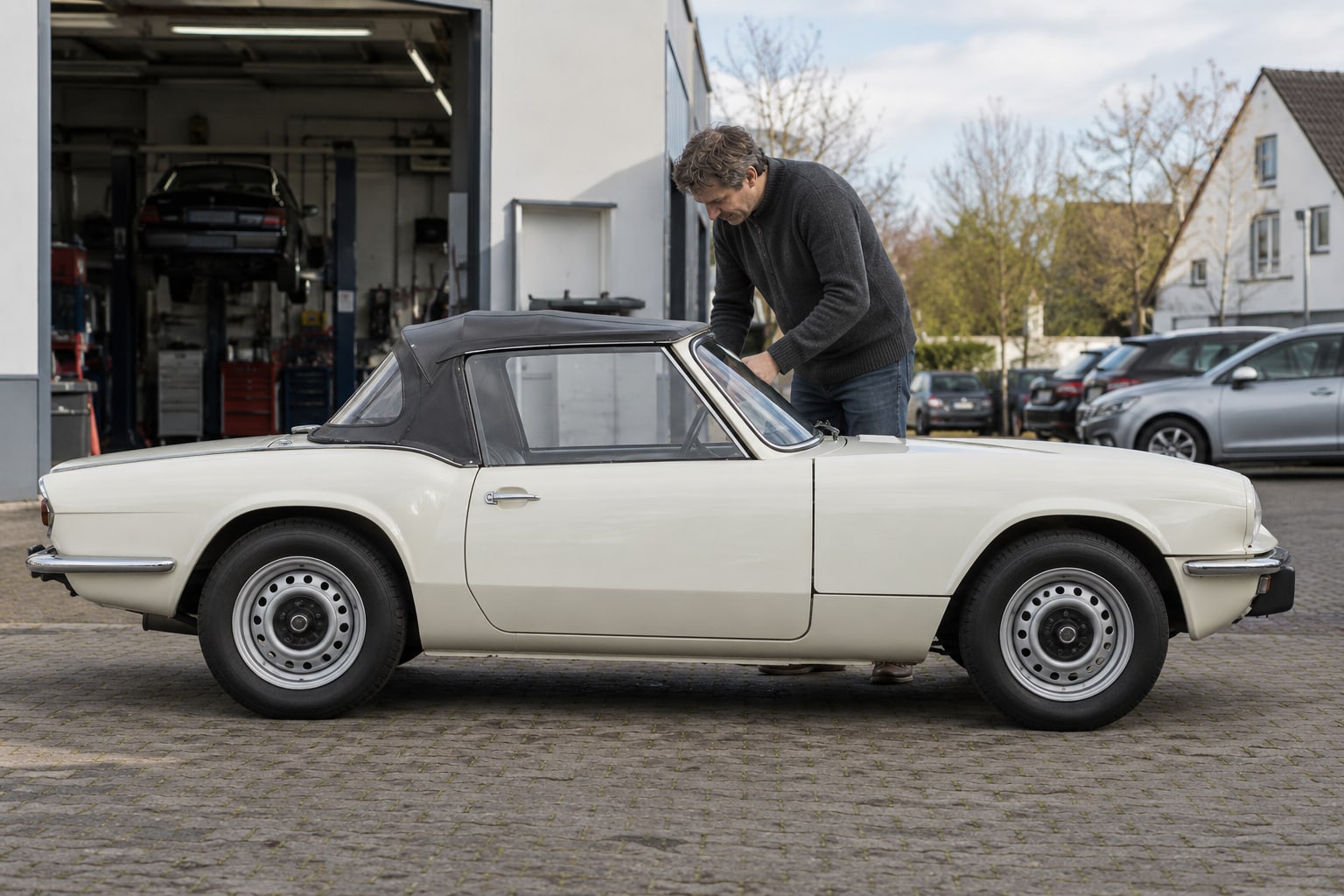 Triumph Spitfire Autoversicherung: Baureihe und zeitweise abgenommene Dachteile in die Vertragsprüfung einbeziehen. Weißer offener klassischer Roadster mit Mann vor einer Werkstatt: Triumph Spitfire Kfz-Versicherung