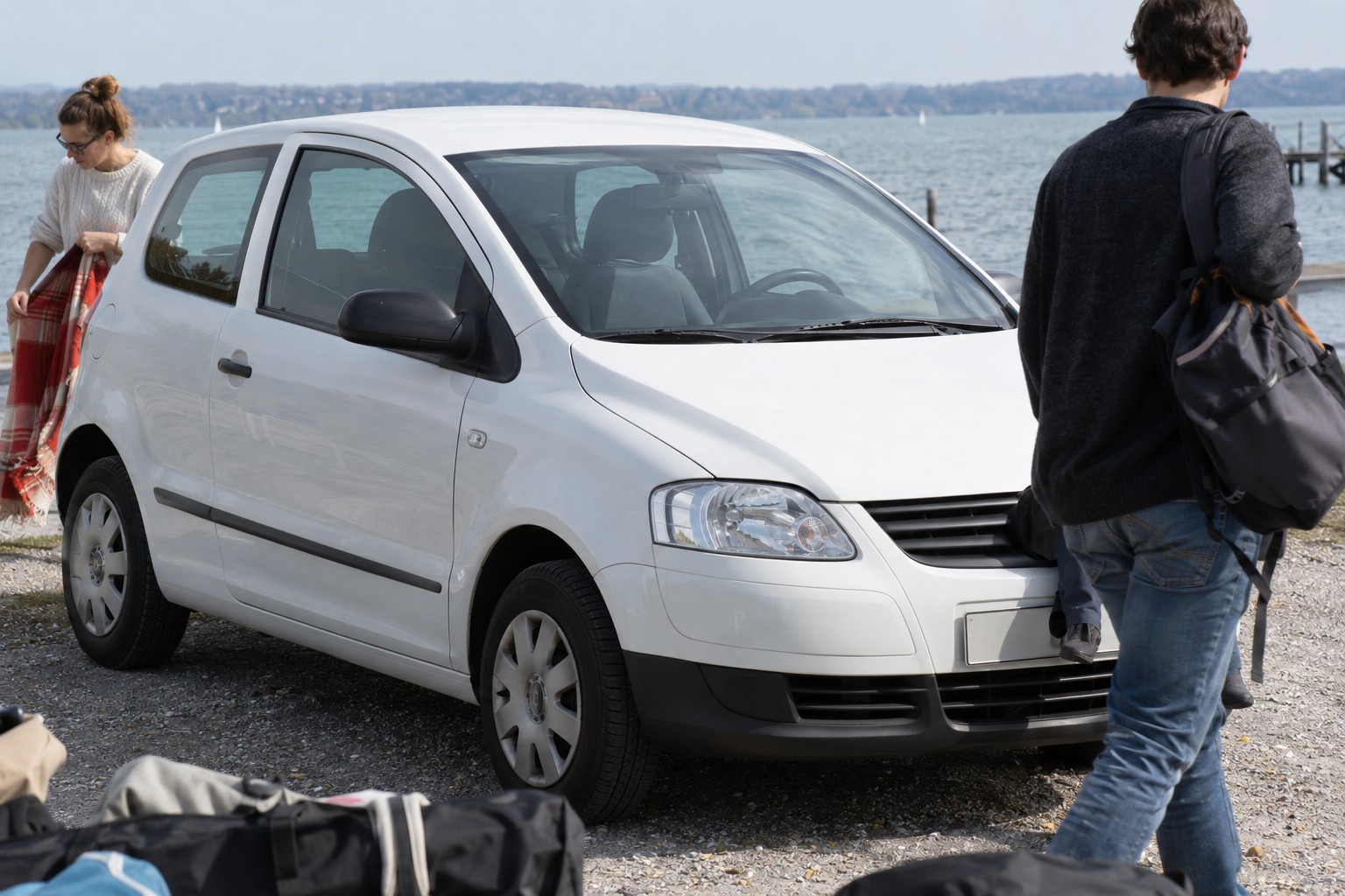 VW Fox: Ein Fox als älterer Zweitwagen sollte nach Restwert und vorgesehenen Fahrern versichert werden. Weißer VW Fox auf einem Seeparkplatz neben einer Person mit Picknickdecke
