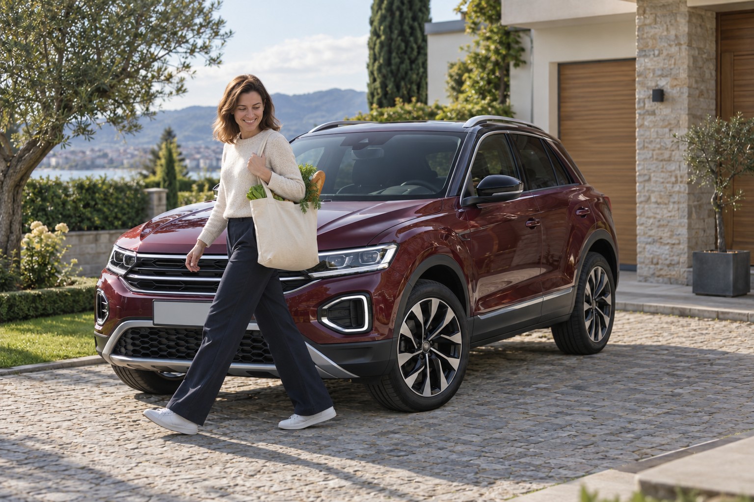 Bordeauxroter VW T-Roc auf einer Hauseinfahrt; eine Frau geht mit einem Einkaufsbeutel vor dem Fahrzeug vorbei