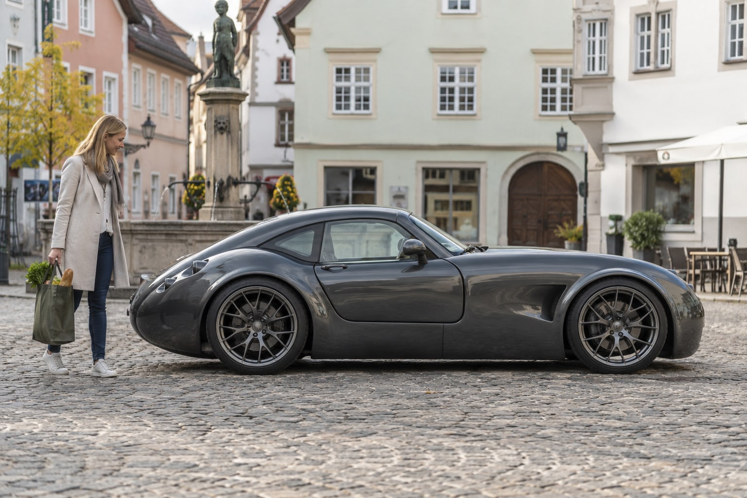 Wiesmann MF5 Autoversicherung: V10 und spätere Biturbomodelle verlangen getrennte Fahrzeug- und Wertangaben. Graues flaches Sportcoupé mit Frau auf einem Altstadtplatz: Wiesmann MF5 Kfz-Versicherung